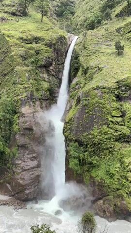 A tall waterfall cascades down a moss-covered cliff face into a churning river below. The surrounding landscape is dominated by steep, green hillsides dotted with trees. The water is white and frothy as it plunges, creating a mist at the base. The rocks of the cliff are dark and appear wet. The overall impression is one of raw, natural beauty and power.