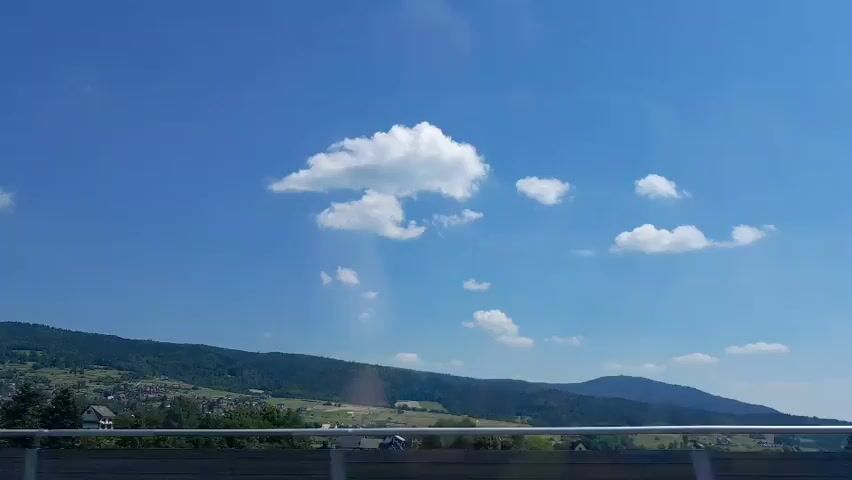 The video shows a view from a moving vehicle. In the foreground, a dark railing is visible. Beyond the railing, a landscape of green hills dotted with houses stretches into the distance. The sky is a clear blue with scattered white clouds. The camera pans slightly to the right, capturing more of the scenery.