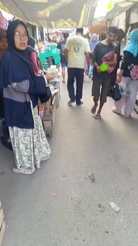 This is a video of a busy marketplace. In the foreground, a person wearing a hijab and a patterned skirt walks away from the camera. To the right, a person in a black shirt and shorts walks away, holding a green object. Further back, several other people are visible, some browsing stalls and others walking through the crowded path. The stalls appear to be selling food items, and there are various goods displayed. The ground is paved, and there are awnings overhead, suggesting an outdoor market setting. The video is somewhat blurry and shaky, indicating it was likely recorded while walking.