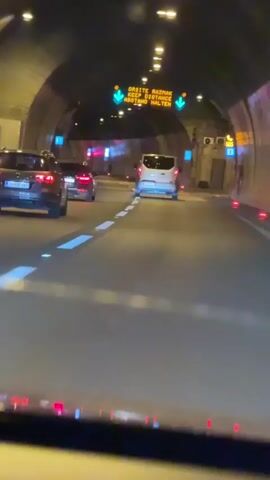 The video shows a view from inside a vehicle driving through a tunnel at night. Several cars are visible ahead, traveling in the same direction. An illuminated sign hangs from the ceiling of the tunnel, displaying a message in multiple languages, including "KEEP DISTANCE," along with directional arrows. The tunnel walls are illuminated by lights, and the road surface is marked with white lines. The overall impression is of traffic moving through a well-lit, enclosed roadway.