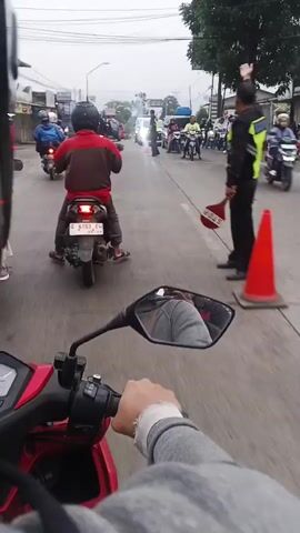 The video shows a street scene with several motorcycles and vehicles. A person on a motorcycle is filming the scene with a forward-facing camera, showing the motorcycle's handlebars and a mirror reflecting their leg. In the distance, there are several other motorcycles, some with riders wearing helmets. There is also an ambulance and what appears to be a traffic police officer directing traffic. The officer is wearing a high-visibility vest and holding a red and white traffic sign. The weather appears to be overcast.