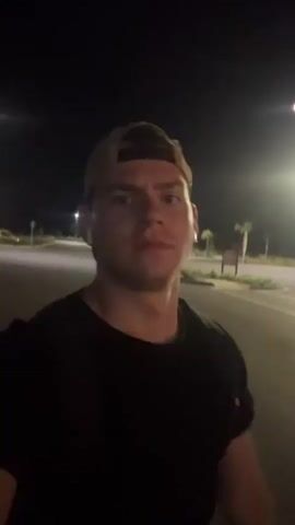 The video shows a young man wearing a black t-shirt and a baseball cap. He is outside at night, likely in a parking lot or on a street, as evidenced by the streetlights and pavement. He appears to be moving and looking at the camera. At one point, he flexes his arm. He is also wearing wireless earbuds.
