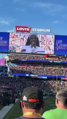 The video shows a wide view of Levi's Stadium, packed with spectators.  At the center of the scene is the stadium's large screen, displaying a performer, standing on a field holding a microphone. In the foreground, a man with a black and orange hat stands with his back to the camera, looking towards the field.  Beside him, partially visible, is another man in a green shirt.  The atmosphere suggests a large, public event, likely a football game given the stadium setting. The screen shows the singer and the SUPER BOWL logo.