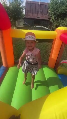 A young girl in a pink t-shirt and striped skirt, wearing a wide-brimmed hat, bounces on a green inflatable surface inside a multi-colored bouncy castle. The bouncy castle has red, orange, and yellow inflatable supports and walls. The girl is smiling and appears to be in motion, with her arms outstretched. Outside the bouncy castle, green grass and trees are visible, along with a building in the background. The sun is shining.