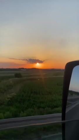This video captures a sunset viewed from the passenger side of a vehicle traveling along a road. The scene is set in a rural landscape with a vast, green field, likely a cornfield, filling the foreground. In the distance, the sun is setting, casting a warm orange glow across the sky and illuminating a line of clouds with rays of light. A single tree stands prominently in the middle ground, silhouetted against the vibrant sky. Beyond the field, industrial structures and buildings are faintly visible on the horizon, suggesting the proximity of a town or industrial area. The sky transitions from a pale blue at the top to a deep orange near the horizon, with a few wispy clouds. The time of day is clearly sunset, evident from the low position of the sun and the rich, warm colors in the sky. The weather appears to be clear and fair, ideal for enjoying a sunset. The view is partially framed by the car's side mirror and a guardrail along the edge of the road, indicating the perspective of someone in transit. There is no visible text within the video. The overall mood is peaceful and serene, characteristic of a beautiful sunset in the countryside. The location context provided is Podłęże, Poland.