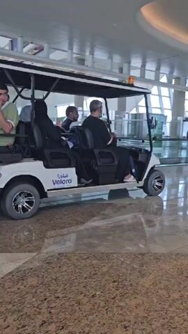 A white golf cart, branded with "Velora" in Arabic, is transporting four people through a modern, spacious airport terminal. The cart has a black roof and sides. The person in the front passenger seat is wearing a black outfit. The driver is also wearing a black outfit. Two passengers are seated in the rear. One passenger, wearing a light green robe with darker green accents, is seated on the left side of the rear seat. A black bag is on the floor behind the rear passenger. The other rear passenger is seated on the right side of the rear seat, wearing a black outfit. The floor of the terminal is made of polished marble, reflecting the overhead lighting. In the background, a moving walkway is visible with several people on it. The architecture of the terminal features large windows, support columns, and curved, illuminated ceiling panels. The lighting appears to be a mix of natural light from the windows and artificial light from the ceiling.