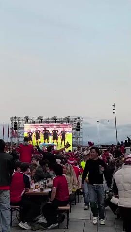 The video shows a crowded outdoor area with many people gathered to watch a large screen displaying a soccer game. Spectators, many wearing red shirts, are seated at tables, some holding drinks and food. In the background, a massive screen mounted on scaffolding shows several soccer players lined up. The sky is overcast. One man in the foreground, wearing a black shirt and jeans, walks past carrying a plastic bag filled with bottles, while a person in a bright yellow vest walks by carrying a similar bag. Some people are standing and cheering, while others are seated and engaged in conversation. The overall atmosphere appears to be one of a public viewing event for a sports match.