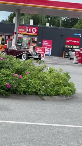 In Laagri alevik, Estonia, a vintage maroon convertible is captured at a gas station. The car, with its distinctive chrome accents and white-walled tires, stands out against the backdrop of the modern Circle K convenience store and Autopesu (car wash) facility. A person is seated in the driver's seat of the convertible.

The scene suggests a bright day, likely late morning or early afternoon, given the lighting and the clear visibility of the surroundings. The presence of gas pumps and the Autopesu sign indicate an active service area. The dense green foliage in the foreground, including some pink flowers, adds a touch of nature to the urban setting.

Visible text includes "Le K" (Circle K logo), "miles+", and "AUTOPESU". There are also Finnish phrases like "Kiire, Lihtne, Joustune" which translates to "Fast, Simple, Flexible," likely advertising services or promotions. The overall impression is a snapshot of a classic car enjoying a modern service stop, hinting at leisure or a special occasion.