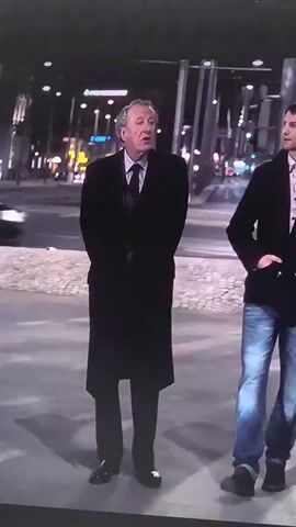 The video shows two men walking outdoors at night, with city lights and buildings visible in the background. The man on the left is older, wearing a black overcoat, suit, and tie. He walks with his hands behind his back. The man on the right is younger, wearing a dark jacket over a light-colored shirt, and blue jeans. He is looking towards the older man and speaking. The scene appears to be a street or sidewalk.
