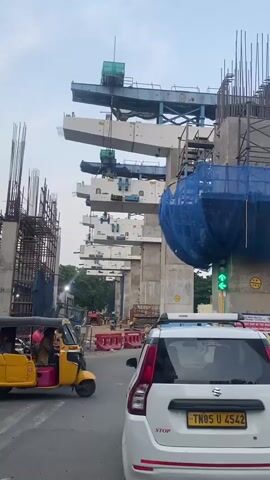 The video shows a street view during the construction of a metro rail line. In the foreground, a white car is stopped at an intersection with a green traffic light. To the left of the car, a yellow auto-rickshaw is also stopped. Behind these vehicles, massive concrete pillars and overhead beams of the metro line construction are visible. Cranes and construction equipment are present in the background, with workers visible on the site. The area appears to be an urban environment with trees and buildings visible in the distance. Red and blue construction barriers are placed along the road. The sky is overcast.