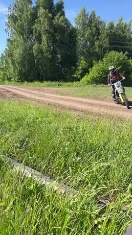A person in a helmet and motocross gear on a green and white dirt bike accelerates rapidly from a patch of tall green grass, driving onto a dirt track that runs diagonally across a field, and then quickly exits the frame to the right. The scene is outdoors on a sunny day with a blue sky, featuring green trees in the background, a distant cell tower, and a solar panel on a pole.