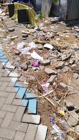 The video pans across a street scene with a paved walkway on the left and a debris-filled area on the right. The walkway is made of interlocking pavers, some of which appear to be chipped or missing. A blue and white painted line runs along the edge of the walkway, bordering the debris. The debris consists of various items including broken bricks, plastic bags, paper, dried leaves, and other miscellaneous trash. In the background, there are structures that appear to be makeshift stalls or buildings, constructed with wood, corrugated metal, and tarps. Some objects like a red chair, a metal grate, and a blue tarp are visible among the debris. The overall impression is one of disarray and neglect.