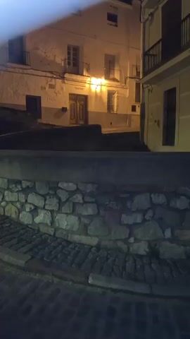 The video shows a nighttime view of a narrow street in a town. In the foreground, there is a low stone wall and cobblestone pavement. Behind the wall, there is a white building with multiple windows and balconies, illuminated by warm light. To the right, another building with balconies is visible. In the background, a tall, illuminated bell tower stands against the dark night sky. The scene is quiet and appears to be uninhabited. The camera is stationary, and the shot is framed from a slightly elevated perspective.