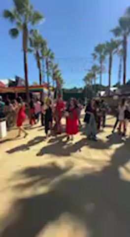 The video captures an outdoor scene on a bright, sunny day, likely in Jerez de la Frontera, Spain, given the context. Numerous people are walking along a wide, unpaved pathway lined with tall palm trees on both sides, suggesting a festive or public event. The sky is clear blue, and strong shadows indicate it's daytime. Many individuals are dressed in what appears to be traditional or festive attire, with several women prominently seen in red and burgundy dresses, some with elaborate ruffles. The atmosphere seems lively and celebratory as people move around, interact, and some appear to be dancing or posing for photos. In the background, there are glimpses of structures that look like market stalls or temporary event tents. No discernible text is visible.