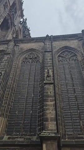 The video pans upwards along the side of a large stone building, likely a cathedral or church. The stone is dark and weathered, with intricate architectural details visible, including pointed arch windows and decorative carvings. Gargoyles or similar decorative figures protrude from the upper portions of the building. The sky above is overcast and grey. In the final frame, the camera angle shifts to show more of the side of the building, including a large arched doorway. A person, wearing a black t-shirt and grey pants, is visible in the lower right corner of the frame, walking away from the building and looking up. The ground in front of the building is paved with cobblestones.