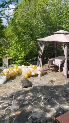 The video shows an outdoor scene on a stone patio. A cluster of white and gold balloons lies on the ground near a black trash bag. In the background, there's a gazebo with a table and chairs set up underneath. The camera pans up to reveal a stone retaining wall with lush green trees and foliage beyond it. In the distance, a panoramic view of a forested landscape extends to a horizon marked by hazy mountains under a partly cloudy sky.