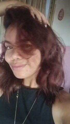 A medium shot captures a woman with auburn hair, her face partially obscured by her hair as she poses for the camera. She wears a black top and a silver necklace with a pendant. The background is blurred, with hints of a pink wall and a light-colored door. The woman pouts her lips, a gesture often associated with taking a selfie, and her arm is raised behind her head, suggesting a playful or flirtatious pose. The lighting is natural and appears to be coming from the side, casting a soft glow on her face.