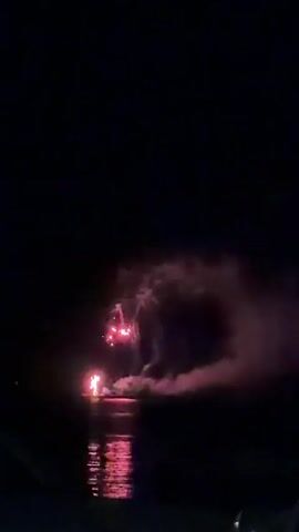 This video shows a nighttime fireworks display over a body of water. The fireworks are primarily red and pink, with some green and white accents. They are reflected in the dark water below. The fireworks are launched from a location on or near the water, possibly a boat or a platform. Smoke from the fireworks drifts across the scene, obscuring some of the lower portion of the display. The foreground is dark and appears to be the edge of the water or a viewing area. The overall mood is festive and celebratory.
