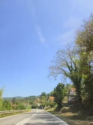A wide shot captures a winding road with white lane markings, leading into a rural village nestled amongst rolling green hills. The sky is a clear, bright blue, with a few wispy clouds visible. Trees with green foliage line the roadside, and several houses with red-tiled roofs are scattered across the landscape. The camera appears to be mounted inside a vehicle, as evidenced by a slight reflection on the upper portion of the screen. The scene is peaceful and serene, suggesting a tranquil drive through the countryside.