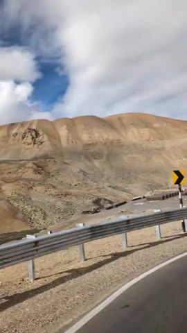 The video captures a roadside view of a mountainous, arid landscape. A guardrail runs along the side of a paved road, which curves to the right. In the distance, a cluster of buildings and structures is visible in a valley. The terrain is characterized by dry, sandy hills with sparse vegetation. Several road signs are present, including a chevron sign indicating a curve, a parking sign, and a circular sign with a red border and a blue symbol. A milestone marker with text is also visible. The sky is partly cloudy, with dramatic formations of white and gray clouds against a blue sky. The overall impression is of a remote, high-altitude region.