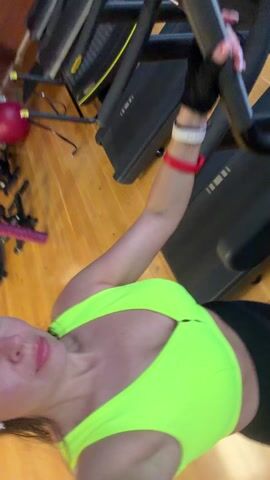 A woman wearing a neon green sports bra and black leggings is on a treadmill at a gym. The camera is oriented towards her face as she walks on the treadmill. Other gym equipment, including additional treadmills and exercise balls, are visible in the background. The gym has wood floors and brick walls with decorative lighting. Another person is visible in the background.