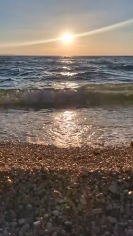This is a vertical, handheld shot of a pebble beach with the ocean and a sunset in the background. The sun is setting over the ocean, casting a golden glow on the water and the pebbles. Waves are gently rolling onto the shore. In the distance, there is a person swimming in the ocean. The sky is a clear blue with some wispy clouds. The overall mood is peaceful and serene, evoking a sense of calm and relaxation. The video appears to have been filmed in Zadar, Croatia, known for its beautiful coastline and sunsets. There is no visible text in the media file.