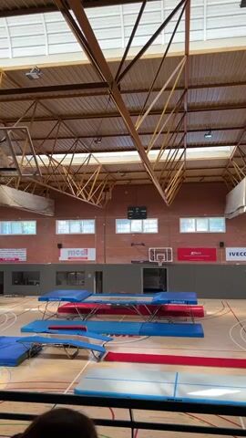 A high-angle, eye-level video shows a high school gymnasium, with multiple trampolines and gym mats laid out on the basketball court. The brick wall at the far end has an electronic scoreboard, a basketball hoop, and various advertisements. The ceiling has exposed wooden trusses and a skylight. No people are visible.