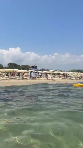 The video shows a sunny day at a crowded beach. In the foreground, the clear, shallow water reveals seaweed on the sandy bottom, with gentle ripples on the surface. Further back, the beach is lined with rows of white umbrellas and lounge chairs, with people scattered among them. A small white building, possibly a concession stand or lifeguard station, is visible on the sand. A yellow kayak floats in the water near the shore, and a red buoy is also present. In the middle ground, two white sailboats with tall triangular sails are navigating the water. The background features a hilly, tree-covered coastline under a clear blue sky with a few white clouds. The scene depicts a leisurely summer day at a popular beach resort.