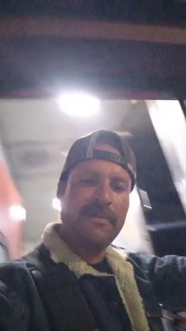 A man in a hat, jacket and mustache stands inside a train and looks towards the camera. He stands with his hand on a vertical, silver bar near the door. The train moves along the tracks at night, next to a paved platform with street lights.
