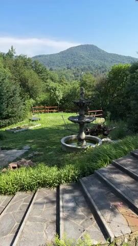 A still image shows a tiered stone fountain in a grassy yard with a stone stairway in the foreground. Beyond the fountain, a wooden fence encloses the yard, and a mountain rises in the distance, covered in trees. A three-headed lamppost stands to the right of the fountain. Lush green trees surround the yard, and the sky is clear and blue.