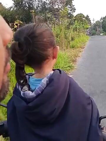 A child, seen from behind, wearing a dark blue hooded jacket over a light blue shirt, sits on a motorcycle. The child has dark hair pulled back into a ponytail. The child points and looks towards the right side of the frame. The motorcycle is traveling on a paved road in a rural area. Lush green grass and trees line the road, with some houses visible in the distance. The sky is overcast.