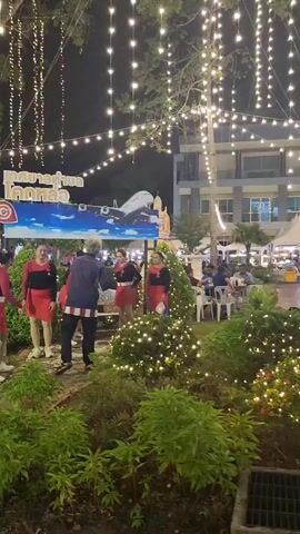 The video shows an outdoor scene at night, illuminated by numerous strings of fairy lights. In the foreground, there are several bushes decorated with more lights. A path made of what appears to be railway ties leads through the bushes. A group of five people, three women in matching red and black outfits with red skirts, and two individuals dressed more casually, are gathered near a sign with Thai writing and an image of an airplane. The women appear to be performing or posing, with two of them holding onto each other. The person in the middle, dressed in a dark jacket with a striped pattern on the sleeves and pants, is facing the women. In the background, there are more trees with lights, tents, tables and chairs with people seated, and a building. Further left, a memorial with a statue, floral arrangements, and a framed picture is visible. There is a black trash bag in the lower left corner of the frame.