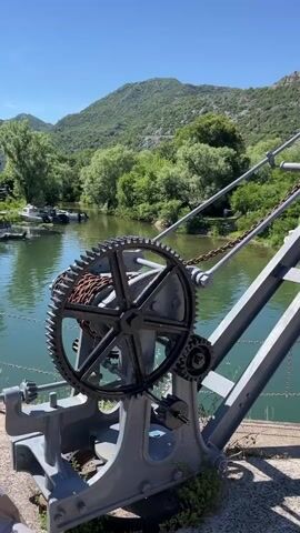 This video shows a close-up of a large metal gear mechanism with a chain attached to it. The gear is part of a crane-like structure. In the background, there is a body of water, likely a river or lake, with lush green trees and hills on the opposite bank. Several small boats are visible on the water, some with people on or near them. The sky is clear and blue, indicating a sunny day. The camera pans slightly, offering a wider view of the scenic environment.