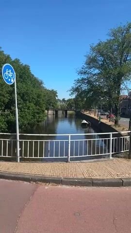 The video shows a canal with a bridge in the distance. There is a bicycle path beside the canal with a sign indicating a bicycle route. A few people are seen riding bicycles. The sky is clear and blue. The sun is shining. There are trees on both sides of the canal. There is also a traffic light at an intersection.