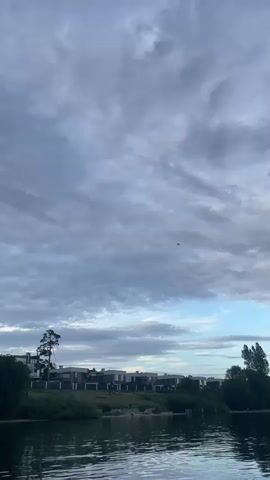 A drone flies in the sky above a body of water and a row of houses. The sky is filled with clouds.