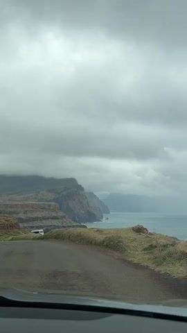 The video shows a view from inside a vehicle, likely a car, driving on a narrow asphalt road. The road runs parallel to a rugged coastline. The landscape is dominated by steep, rocky cliffs and layered rock formations descending to the sea. The cliffs are covered in some areas with grassy vegetation.  The sky is overcast with heavy grey clouds, contributing to a gloomy atmosphere. The sea stretches out to the horizon, with waves crashing against the rocks along the shore. The vehicle appears to be moving forward, offering a continuous view of the coastal scenery as it drives along the road. Music is playing in the background.