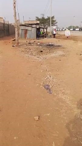 The video shows a barren roadside area. There is a corrugated metal shack to the left, with some people visible inside. In the middle of the foreground, there is a patch of charred ground, suggesting a fire recently took place. The ground is mostly dirt and gravel, with some scattered debris and a discarded piece of cloth. In the background, across the road, there's a signpost and some indistinct vehicles. A person in a white robe is standing near the signpost. The video is shot in daylight, with a slightly hazy atmosphere. The camera appears to be stationary.