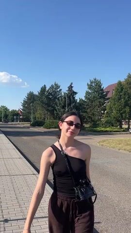 A young woman with dark hair, wearing sunglasses, a black one-shoulder top, and brown pants, walks along a paved sidewalk. She carries a small black crossbody bag. The woman is on a sidewalk next to a road, with trees and buildings in the background. The sky is blue with a few white clouds, suggesting a clear, sunny day. The scene appears to be in a suburban or rural area, possibly in Chotěšov, Czechia. The woman looks at the camera and smiles faintly, appearing relaxed and happy. The video is taken during the daytime. There is no visible text in the media.
