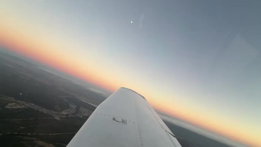 The video shows a view from inside an airplane, looking out over the wing. The sun is setting, casting a gradient of orange, pink, and yellow across the horizon. The landscape below is a mix of trees and buildings, with a body of water visible in the distance. A faint crescent moon is visible in the sky. The airplane appears to be in a gentle bank.