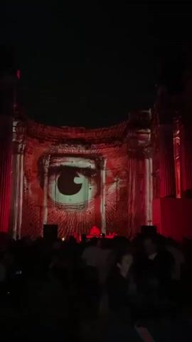 A crowd of people stands in silhouette at the bottom of the frame, facing a large, ruined structure illuminated by dramatic red and white projections. The projections cast an image of a large, stylized eye with swirling patterns onto the ancient stone facade. A small stage with speakers and DJ equipment is visible in the center of the crowd, with a person on it. The overall atmosphere is dark and electric, suggesting an outdoor concert or artistic event. The video is short and transitions quickly between different visual elements of the projection and the crowd.