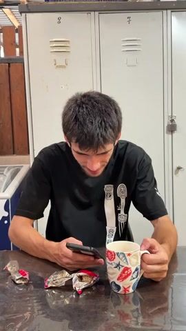 A young man with dark hair is sitting at a table, looking at his smartphone. He is holding a white mug with a seashell pattern in his left hand. In front of him on the table are three crumpled candy wrappers and the smartphone. He is wearing a black t-shirt with a graphic of a knife and fork on it. The background appears to be a locker room with white lockers.