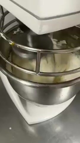 A white commercial-grade stand mixer is actively mixing ingredients in a stainless steel bowl, enclosed by a safety cage. The ingredients appear to be a light yellow or cream color. The mixer is placed on a stainless steel countertop.