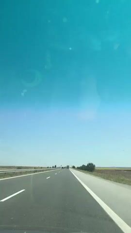 This video shows a view from inside a vehicle traveling on a highway. The perspective is forward-facing, looking out through the windshield. The highway has multiple lanes and a white line marking the edge of the road. On the left side of the highway, there is a barrier and a flat, open landscape. On the right side, there are trees and a field. The sky is a clear blue, with some reflections on the windshield, indicating a sunny day. There are a few other vehicles visible on the road in the distance. The video is short and shows a continuous forward motion.