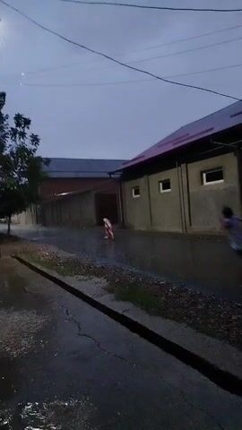 The video shows a street scene at dusk or during a storm. Two children are visible running on a wet asphalt road. One child, wearing a light-colored shirt and dark shorts, is running away from the camera towards a building in the distance. Another child, wearing a dark shirt and light-colored shorts, is running towards the camera on the right side of the road, also appearing to be in motion. The road is wet and reflects the dim light. Buildings with tiled roofs and windows line the right side of the street. A lamppost is visible on the left side of the frame, and trees are present further down the street. The sky is overcast and appears to be dark, suggesting rain or the aftermath of a storm. The overall impression is a brief glimpse of children playing outdoors in wet conditions.