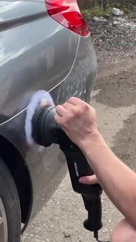 A person's hands are holding a black rotary polisher with a white polishing pad, applying it to the rear quarter panel of a gray car. The polisher is in motion, creating a fuzzy white circular effect on the car's surface. The car has a red taillight visible at the top right of the frame. The tire and part of the wheel rim are visible on the left. The ground appears to be gravelly or dirt. The context suggests car detailing or paint correction.