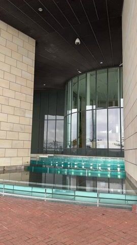 The video shows an exterior view of a modern building with tan brick walls and a large, curved glass window. The building is flanked by brick columns and features a reflective pool in front of the window, with water levels stepping up toward the entrance. The ground in front of the pool is paved with red bricks. A security camera is visible above the glass entrance. The sky is overcast, and reflections of nearby trees can be seen in the glass windows. People are audible in the background, suggesting a public area.