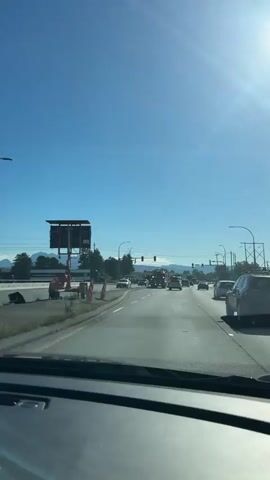 The video shows a view from a vehicle traveling on a multi-lane road on a sunny day. In the distance, there is a tow truck with its hazard lights flashing, positioned in the middle lane. Several other vehicles are visible on the road, and there are traffic lights overhead. To the left of the road, there is a large billboard structure. Further down the road, on the right side, more traffic cones and what appears to be a fire truck or emergency vehicle are present. The sky is clear blue with the sun shining brightly, creating lens flare. In the background, mountains are visible. The video appears to be recorded from a dashcam or a dashboard mount.