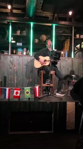 A man is sitting on a stool on a stage playing an acoustic guitar and singing. He is wearing glasses, a black sweatshirt, and black pants. The stage is decorated with flags from France, Canada, and Brazil. Green lights are visible behind the man.
