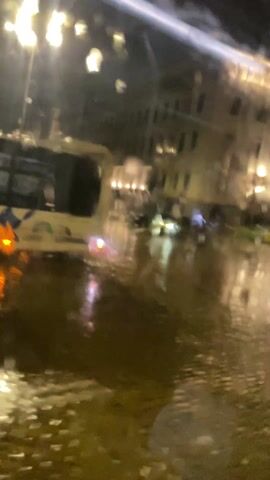 The video depicts a rain-soaked city street at night, viewed from inside a vehicle. The camera is shaky, and the view is somewhat obscured by raindrops on the window. In the background, there is a large building illuminated by a series of lights, reflecting in the flooded street. Two taxis are parked along the street, their taillights reflecting in the water. The street itself is covered in a sheen of water, with reflections of streetlights and building lights visible. The overall impression is of a dark, wet, urban environment.