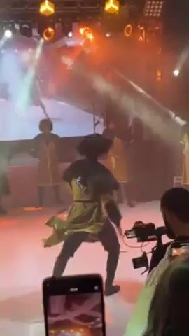 The video shows a group of people performing a traditional dance on a brightly lit stage. Several performers are wearing dark fur hats, long-sleeved outfits with flared skirts, and dark pants. The color of their attire appears to be gold or olive green, with contrasting black accents. One dancer is prominently in the foreground, spinning and leaping across the stage.  Other dancers are visible in the background, in various poses. Stage lighting includes orange and white beams and spotlights. Several audience members are visible near the camera, holding up phones to record the performance.  A professional video camera is also aimed at the stage. The stage appears to be a light color, possibly white or light gray.